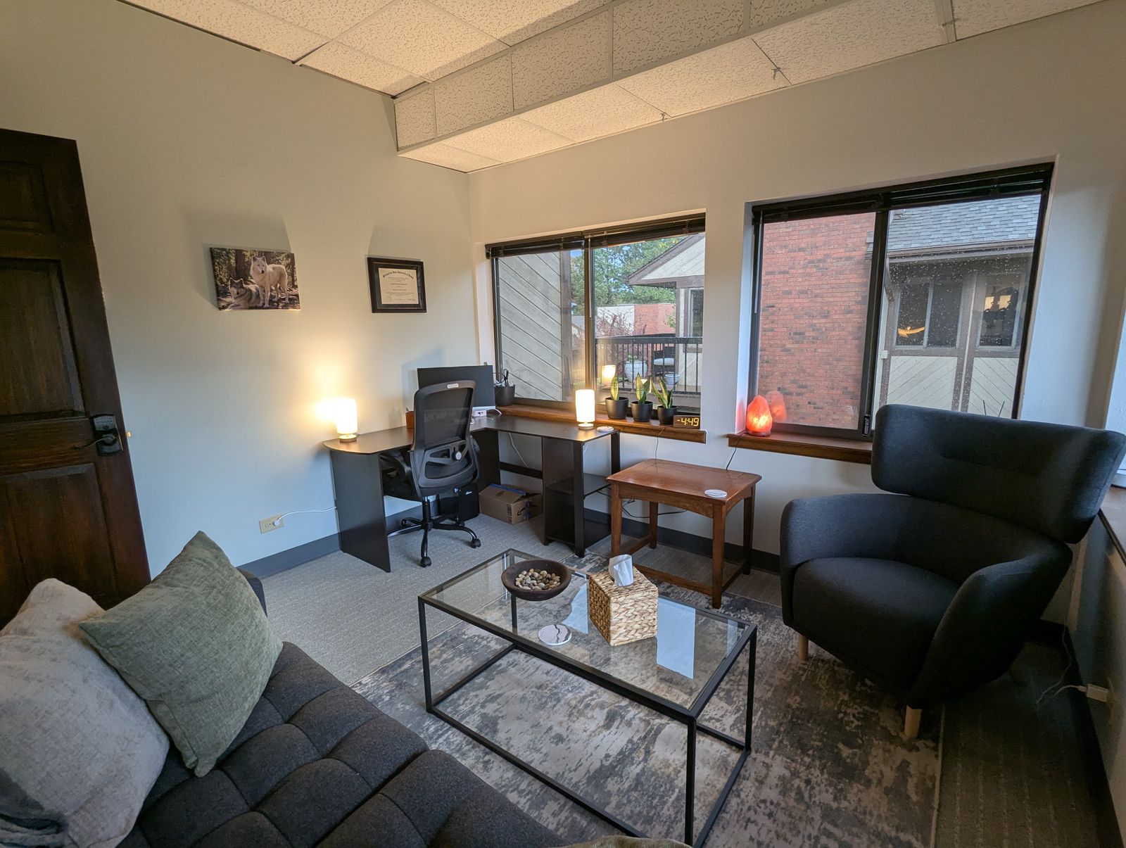 View across the therapy office toward the desk, an armchair, and two windows, with warm lamps on the sills