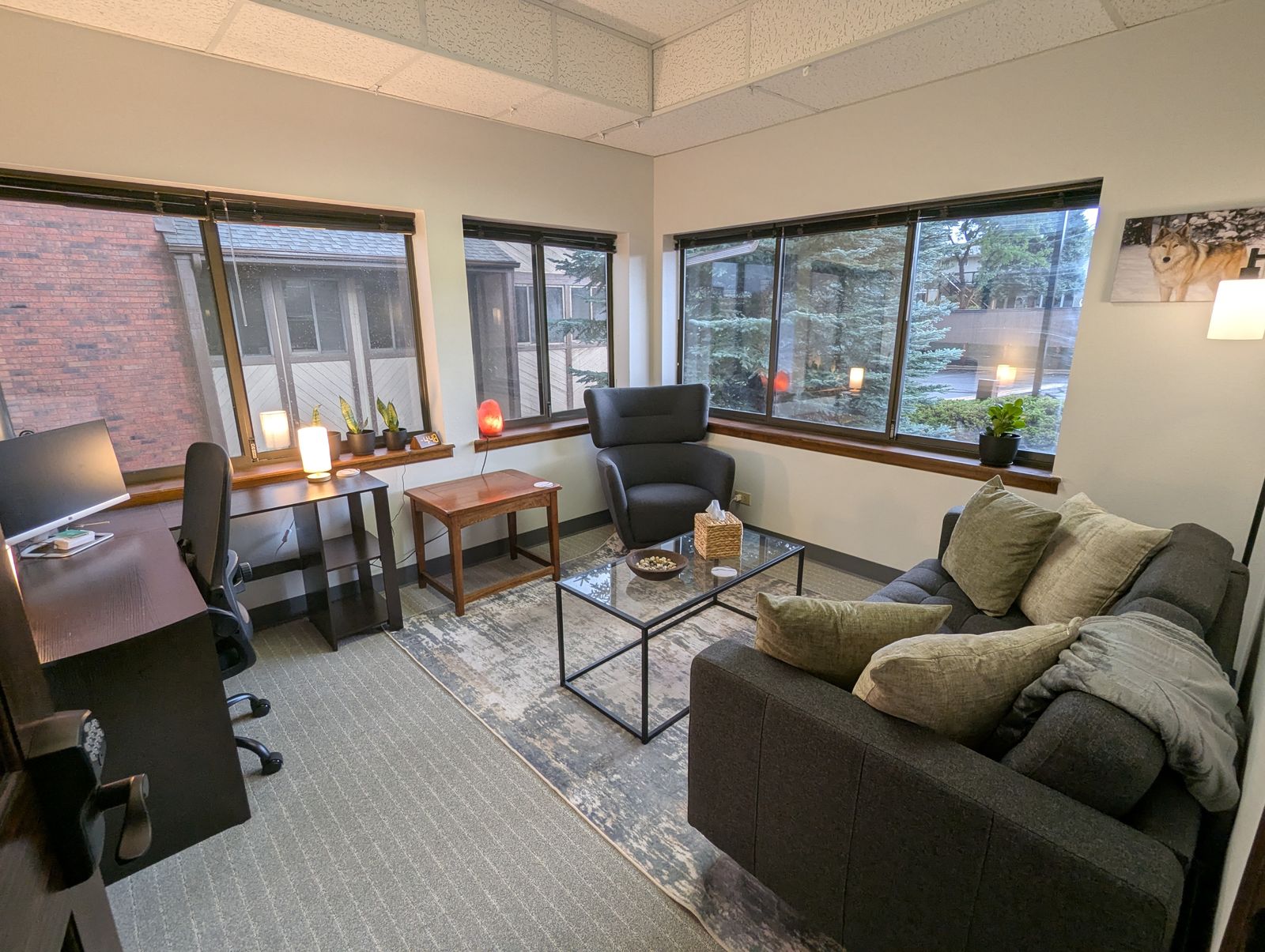 Therapy office with a sofa, armchair, and glass coffee table, lit by corner windows looking out on evergreen trees